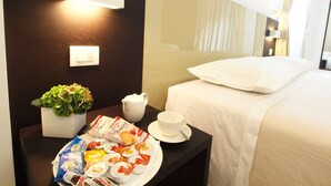 Habitación de hotel impecable con una cama, una mesita de noche con un plato de pasteles, una taza, una jarra y una planta en maceta.
