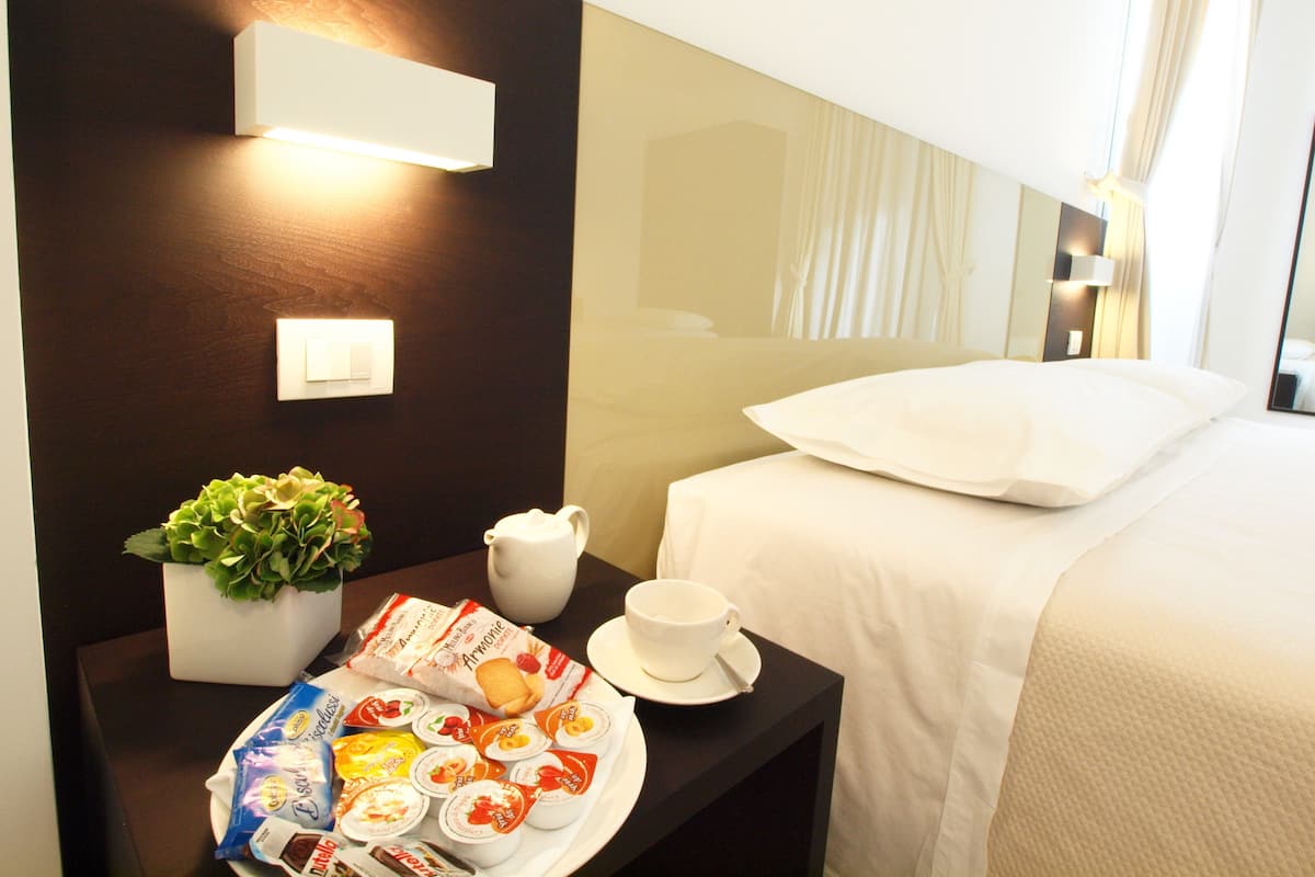 Une chambre d’hôtel bien rangée, avec un lit, une table de chevet sur laquelle se trouve une assiette de pâtisseries, une tasse, un pichet et une plante en pot.