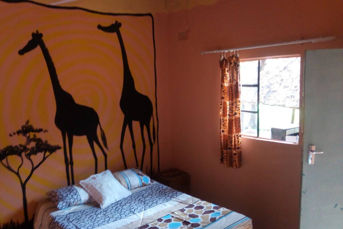 A bedroom with a bed, a window with curtains, and a wall tapestry of giraffes and trees.
