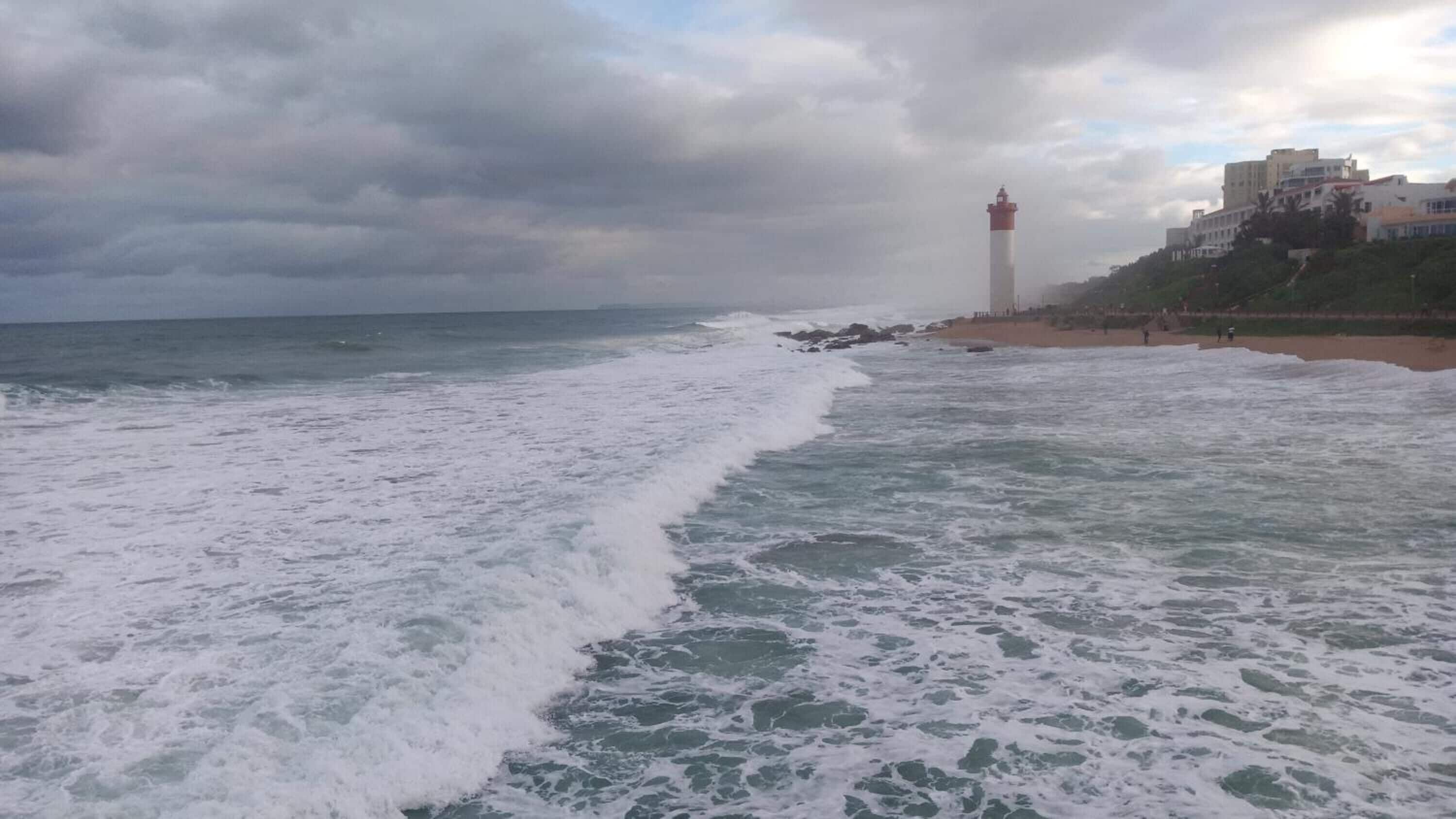Een strand met golven, een vuurtoren en gebouwen op de achtergrond.