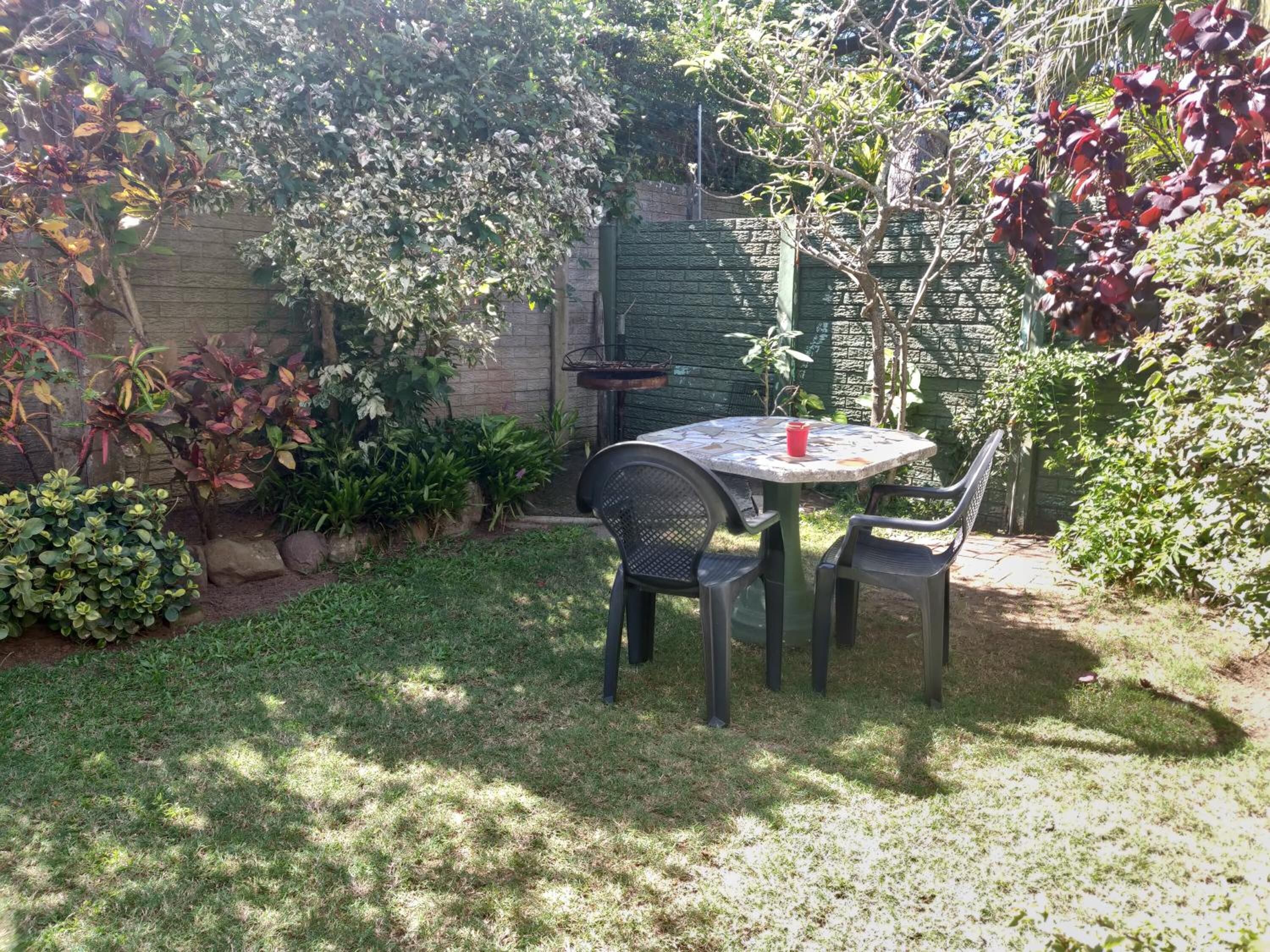 Eine kleine Terrasse im Freien mit einem runden Tisch und zwei Stühlen, umgeben von Pflanzen und einer Backsteinmauer.