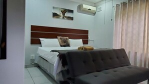 Quarto com cama, cabeceira, mesa de cabeceira, cadeira, cortina e ar condicionado fixado na parede.