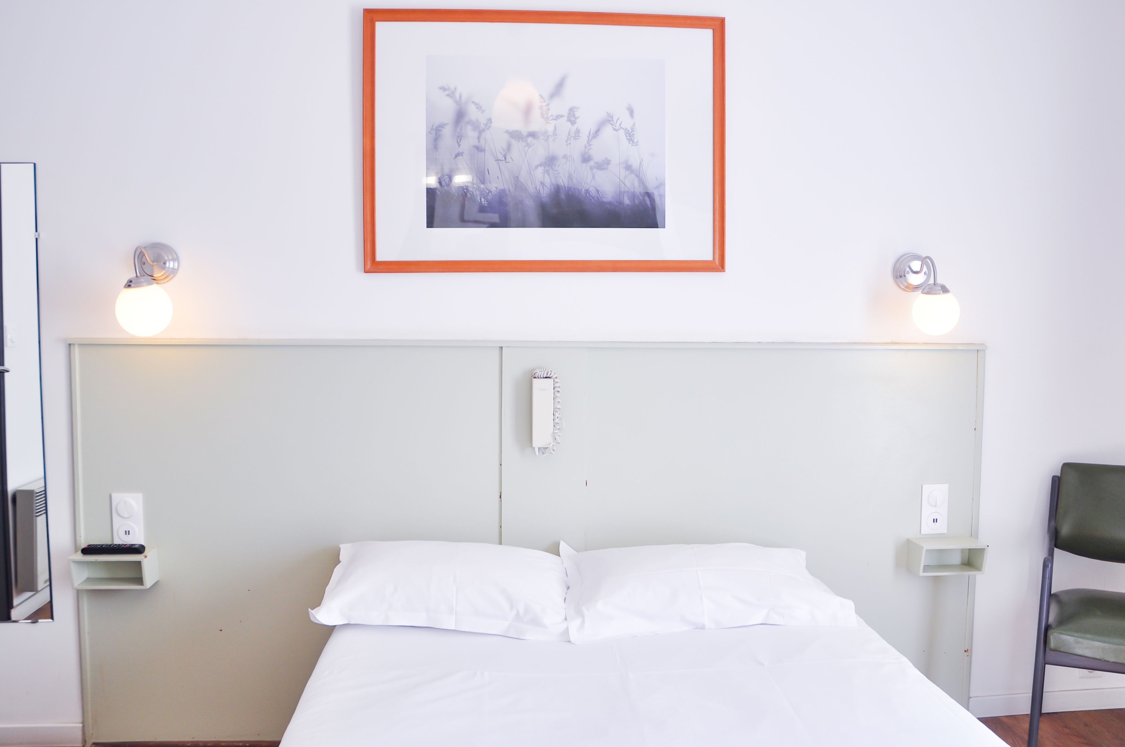 A hotel room with a bed, a framed picture on the wall, bedside lamps, a chair, and a small shelf with a phone.