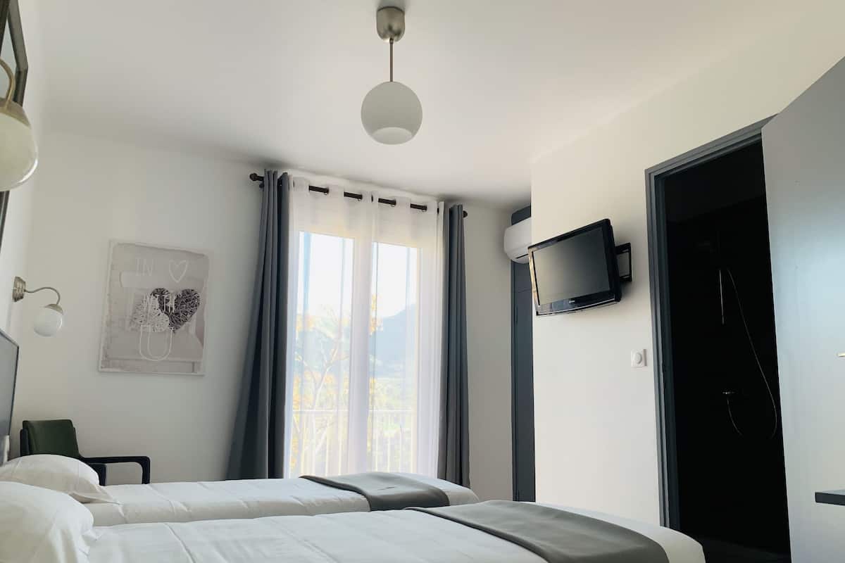 Une chambre d’hôtel avec deux lits, une télévision, une lampe fixée au mur, une fenêtre avec des rideaux et une porte donnant sur un balcon.