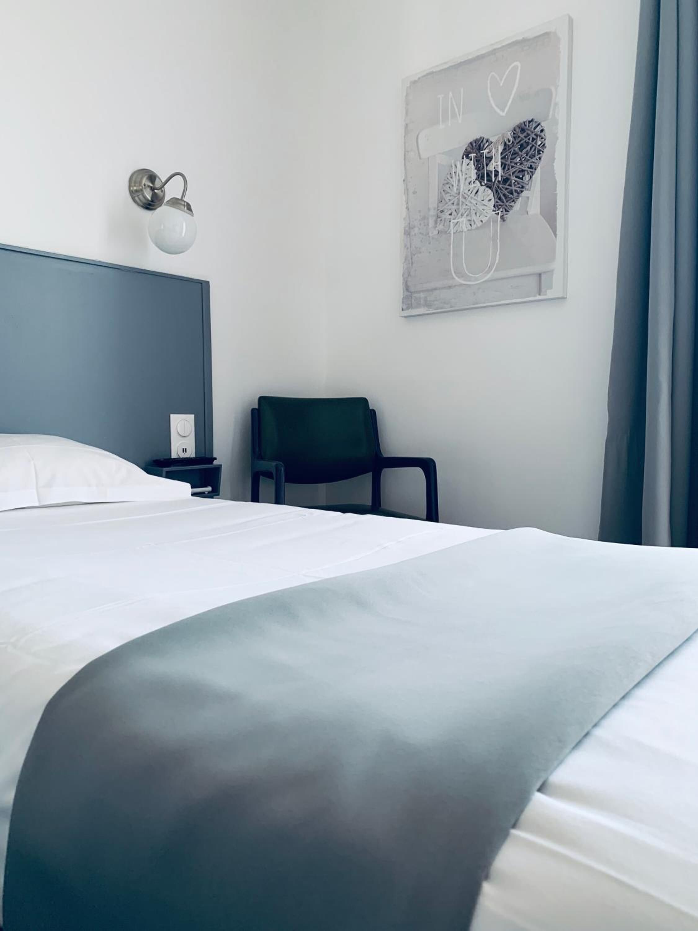 A neatly made bed with a grey headboard, a bedside table with a lamp, and a chair. A wall art piece with a heart and the word "IN" is visible.