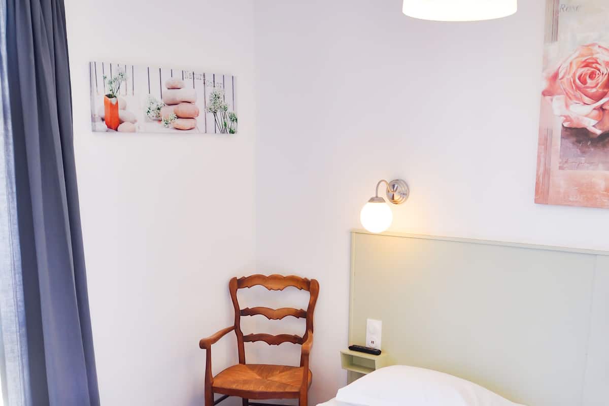 Un lit simple avec du linge de lit blanc, une chaise en bois, une table de chevet avec une lampe et un mur orné d’un tableau encadré.