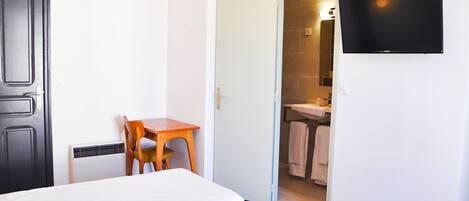 Quarto de hotel com uma cama, uma escrivaninha, uma cadeira, uma televisão e um banheiro visível por uma porta aberta.