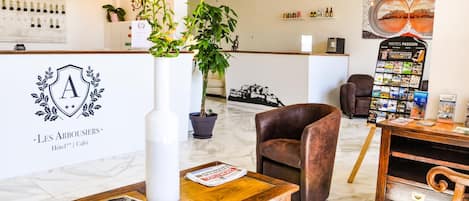 Zona de recepción de hotel con diseño moderno, que incluye un mostrador de recepción, un jarrón grande con plantas, un sillón de cuero marrón y un tablón informativo fijado a la pared.