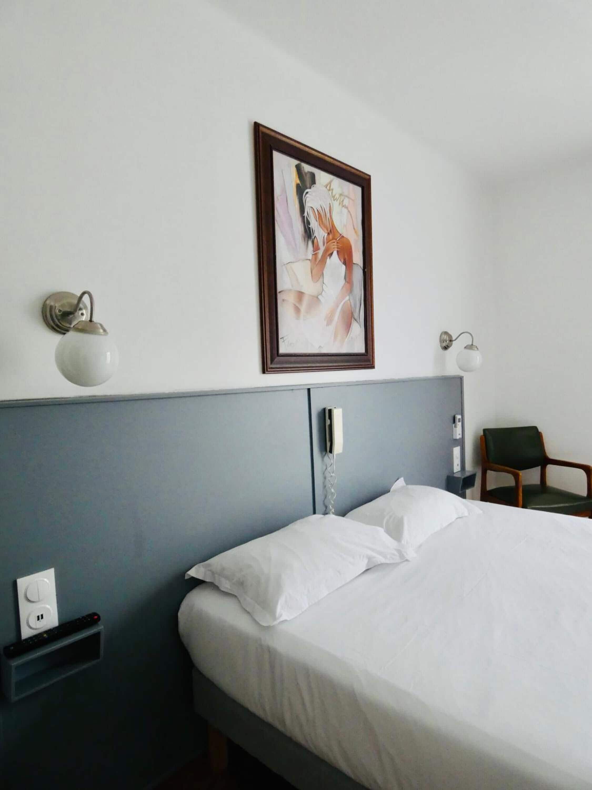 A hotel room with a bed, a wall-mounted telephone, a framed artwork, a bedside table with a lamp, and a chair.