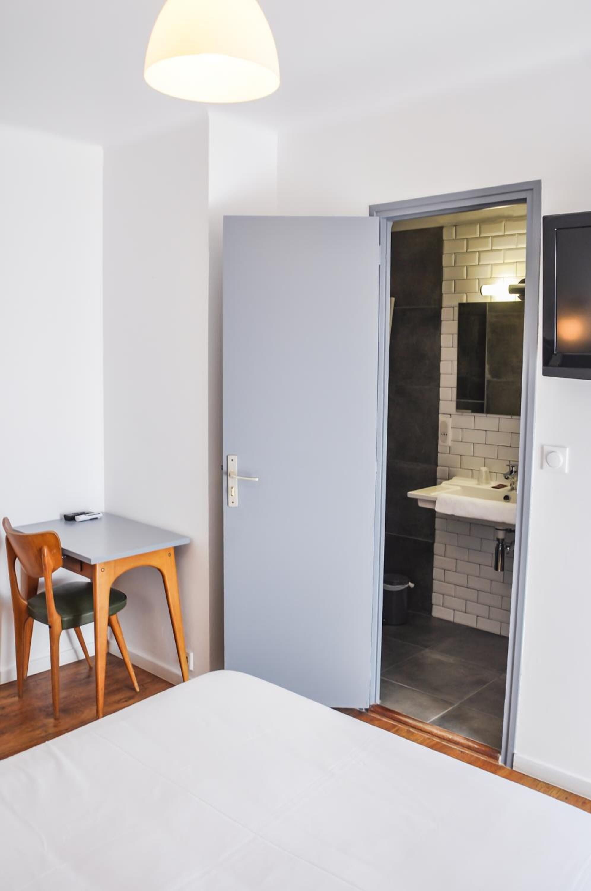 A room with a bed, a table with a chair, a door leading to a bathroom with a sink and mirror.