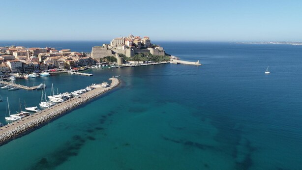 Vista aérea de un pueblo costero con un castillo destacado, un puerto deportivo y aguas azules cristalinas.