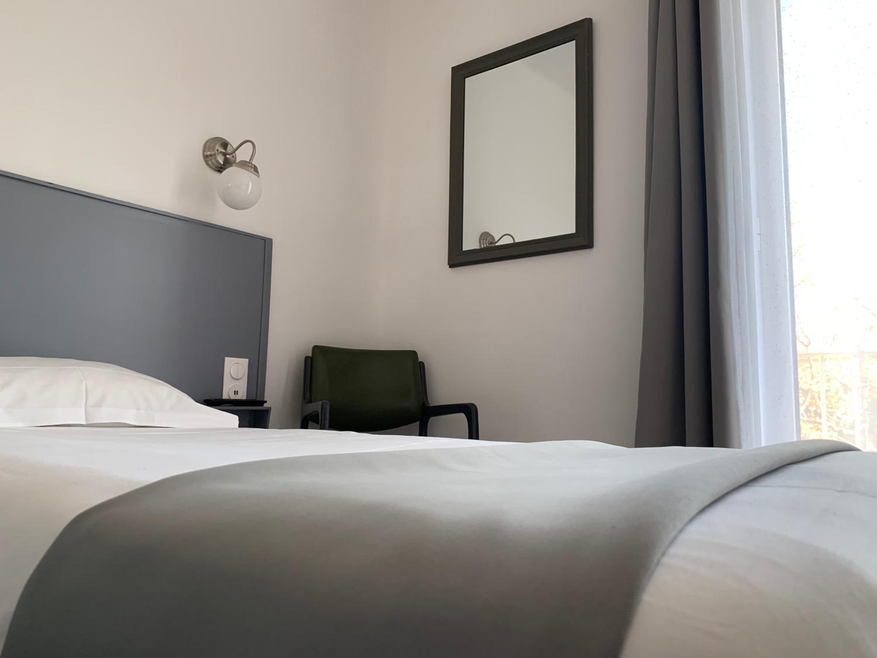 A hotel room with a bed, a chair, a nightstand, a wall-mounted light, and a mirror.