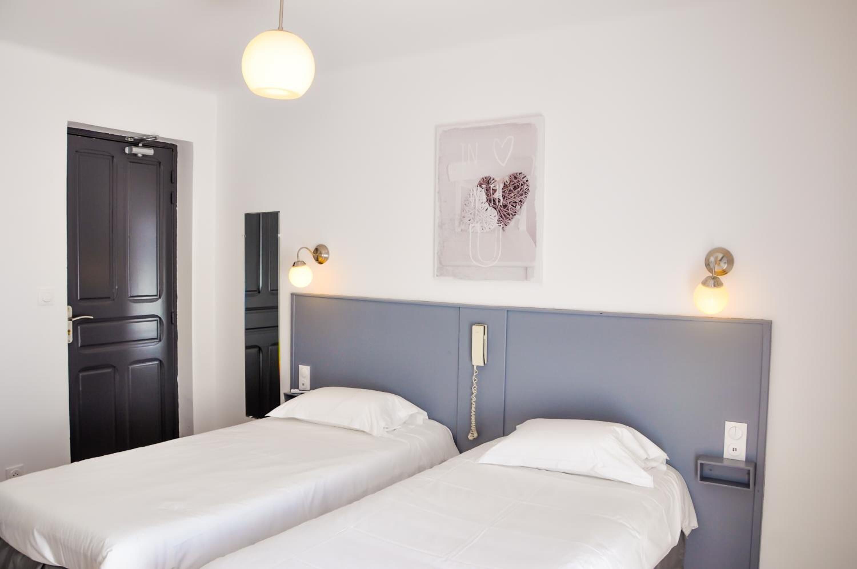 A hotel room with two single beds, a dark door, a light switch, and a wall-mounted phone.