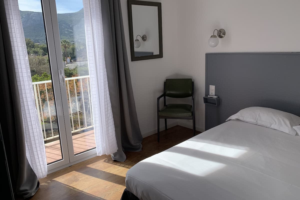 Une chambre à coucher comprenant un lit, une chaise, une table de chevet, un miroir et une vue sur un balcon avec une montagne en arrière-plan.