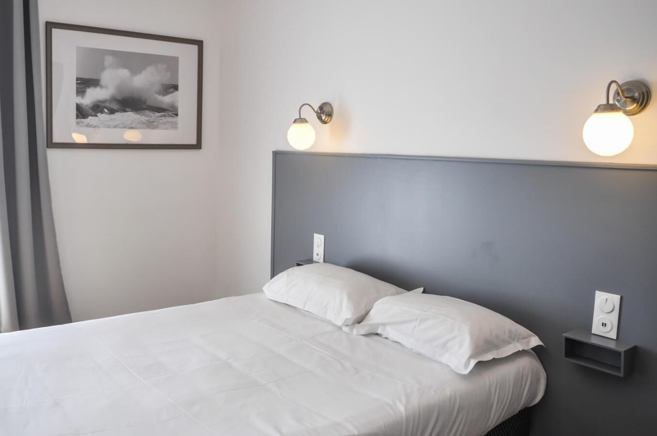 Un lit bien fait, avec du linge de lit blanc, dans une chambre dont la tête de lit est grise. Deux appliques murales sont fixées de part et d’autre de la tête de lit.