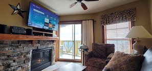 Una sala de estar con chimenea de piedra, un televisor de pantalla plana montado en la pared, un ventilador de techo y vistas al exterior a través de amplios ventanales.
