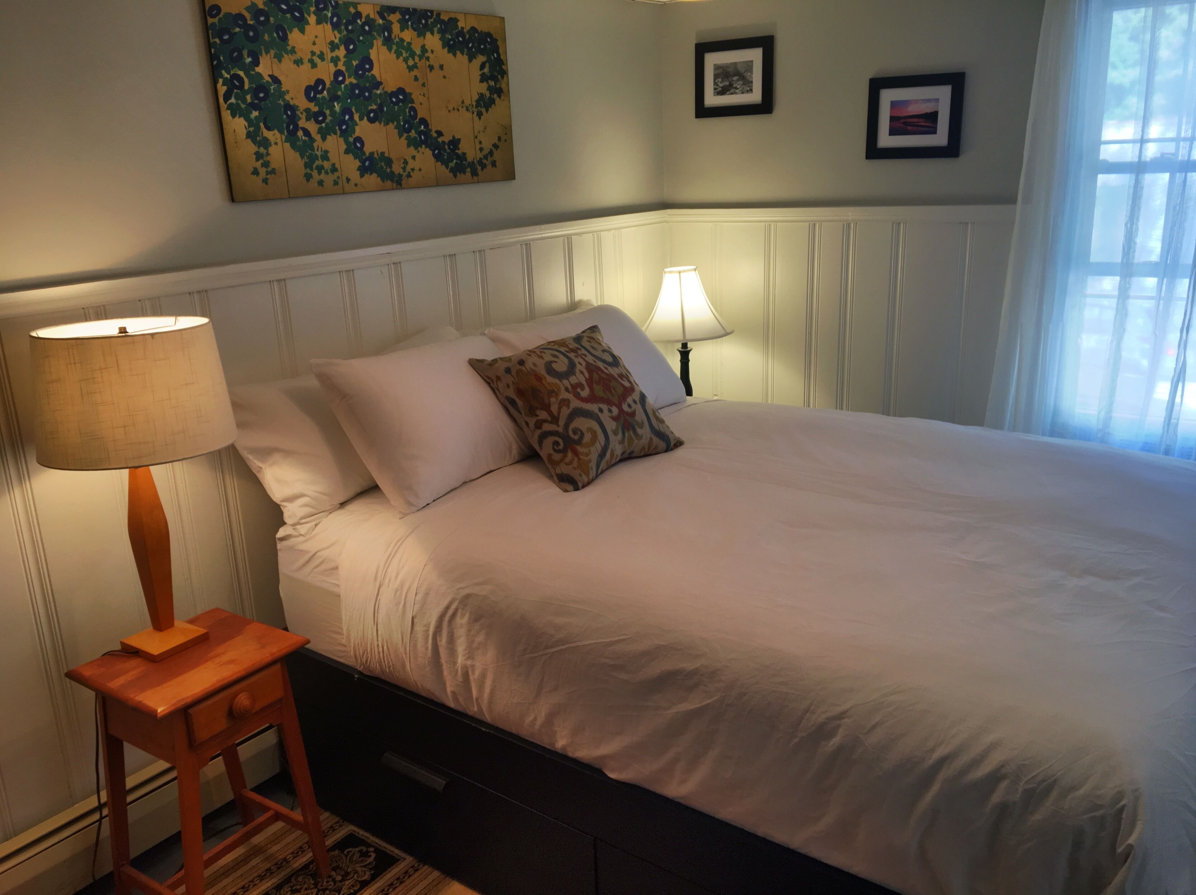 A neatly made bed with white linens, a wooden nightstand with a lamp, and a painting on the wall.