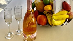 Une table avec une bouteille de vin, deux verres et un panier de fruits contenant des bananes, des pommes et des oranges, donnant sur une piscine et offrant une vue sur l’océan.