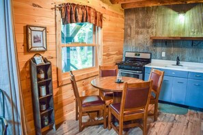 A wooden cabin interior with a dining table, chairs, a stove, and a window with a view of trees.