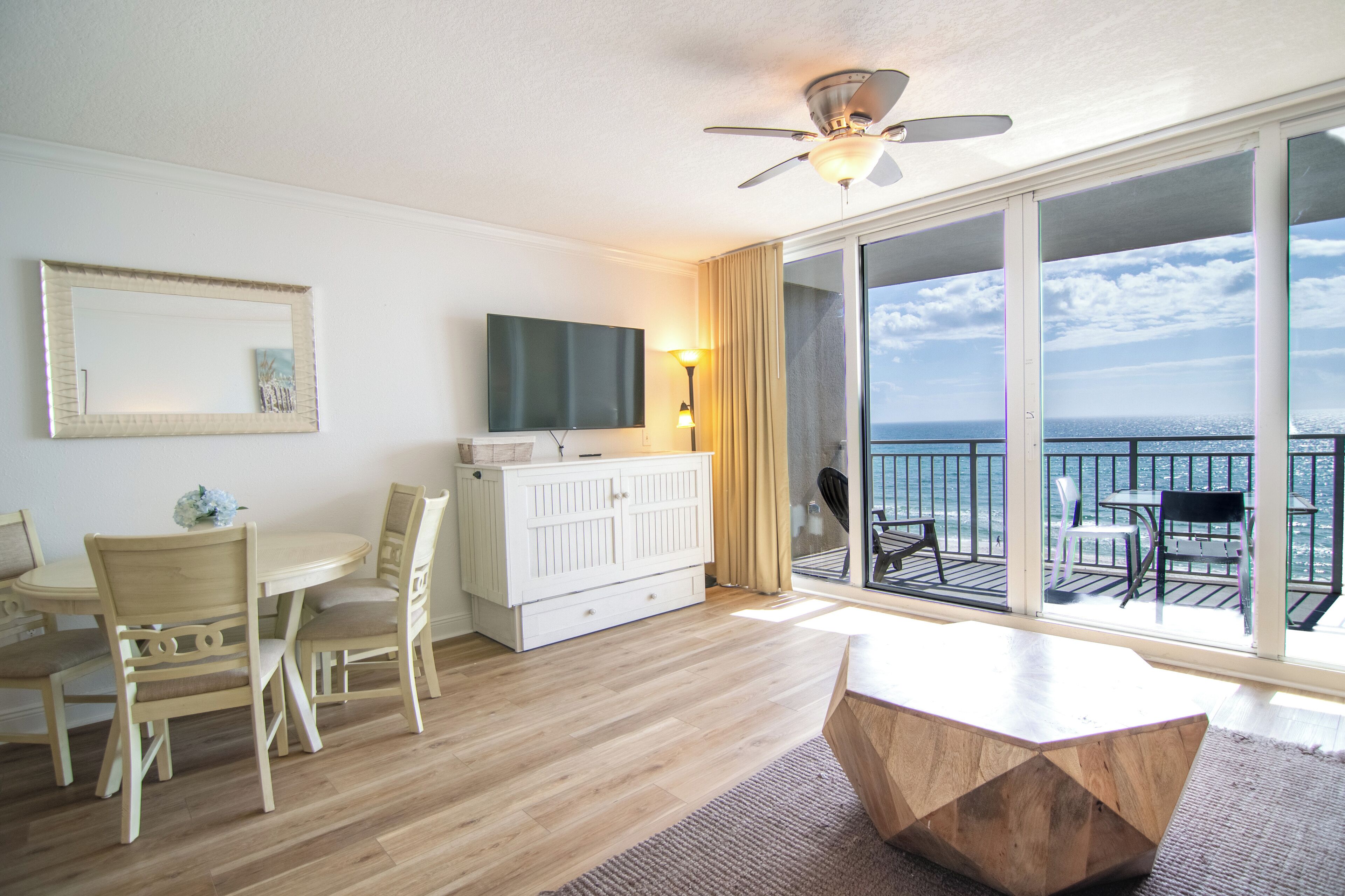 A hotel room with a balcony, a dining area with chairs and a table, a TV mounted on the wall, and a ceiling fan.