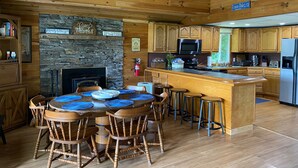 A wooden cabin interior with a stone fireplace, wooden dining table with blue placemats, and a kitchen with wooden cabinets and a stainless steel refrigerator. - Cozy Lakefront Getaway (Union Dale)