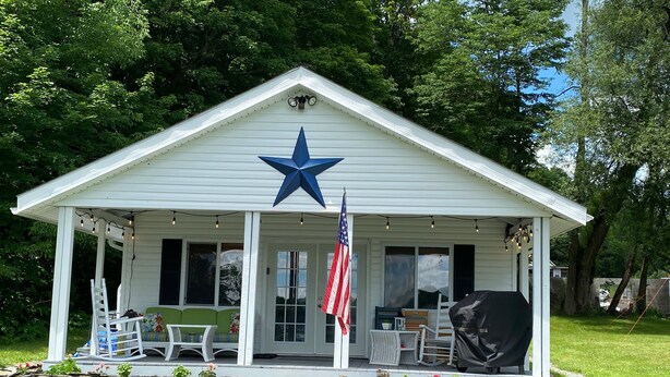 A white building with a blue star on the roof, a covered porch with outdoor seating, and an American flag. - Cozy Lakefront Getaway (Union Dale)