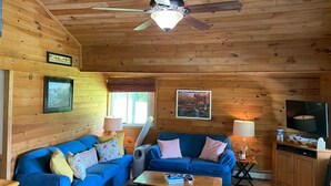 A cozy cabin-style room with two blue sofas, a ceiling fan, a television, and framed pictures on the walls. - Cozy Lakefront Getaway (Union Dale)