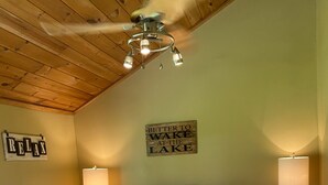 A bedroom with a sloped ceiling, a bed with a floral bedspread, two bedside lamps, a ceiling fan, and a wall sign that reads "Better to wake at the lake." - Cozy Lakefront Getaway (Union Dale)