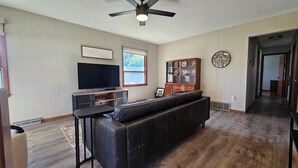 Una sala de estar con un sofá de cuero oscuro, un televisor de pantalla plana, un mueble de madera con platos decorativos y un reloj de pared.