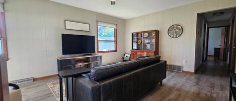 A living room with a dark leather sofa, a flat-screen TV, a wooden cabinet with glass doors, and a decorative clock on the wall.