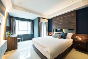 Une chambre moderne avec un grand lit, un bureau et une chaise. On y trouve une fenêtre avec des rideaux, un appareil de climatisation et une table de chevet avec une lampe.