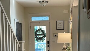 Un pasillo con suelo de madera, una puerta blanca adornada con un adorno navideño, una escalera con barandillas blancas y una mesita con una lámpara y objetos decorativos.