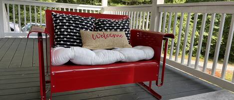 A red bench with white cushions and a 'Welcome' pillow on a wooden deck with white railings.