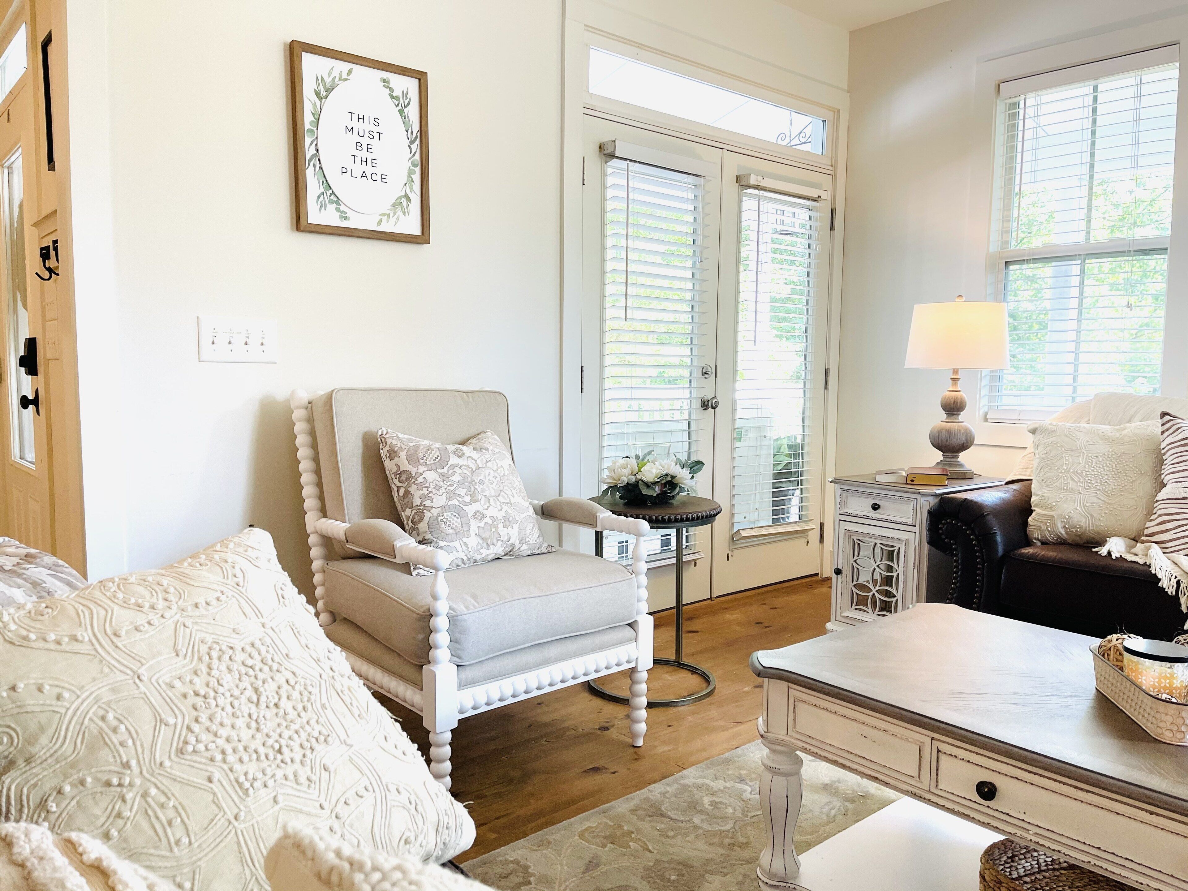 A room with a white armchair, a patterned sofa, a small table with a lamp, and a framed quote on the wall.