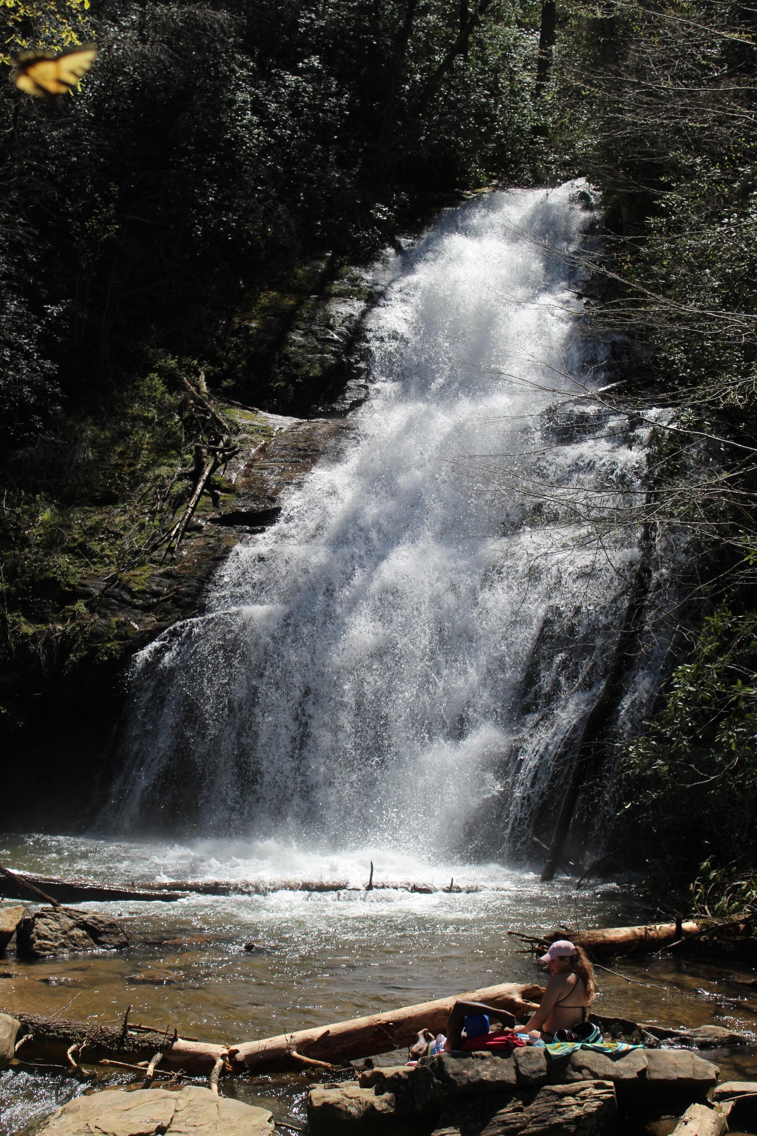 A waterfall in a forested area with a person sitting on rocks near the water.