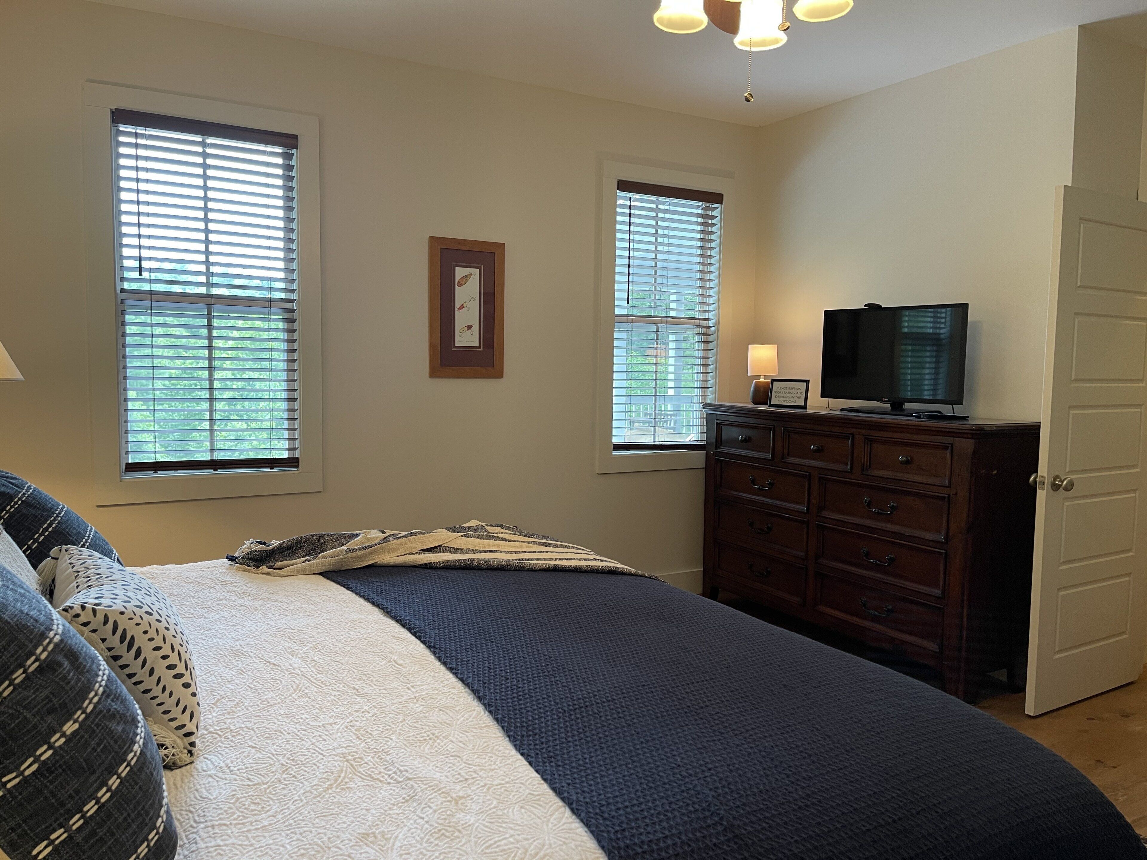 Un dormitorio con cama, cómoda, televisor y dos ventanas.