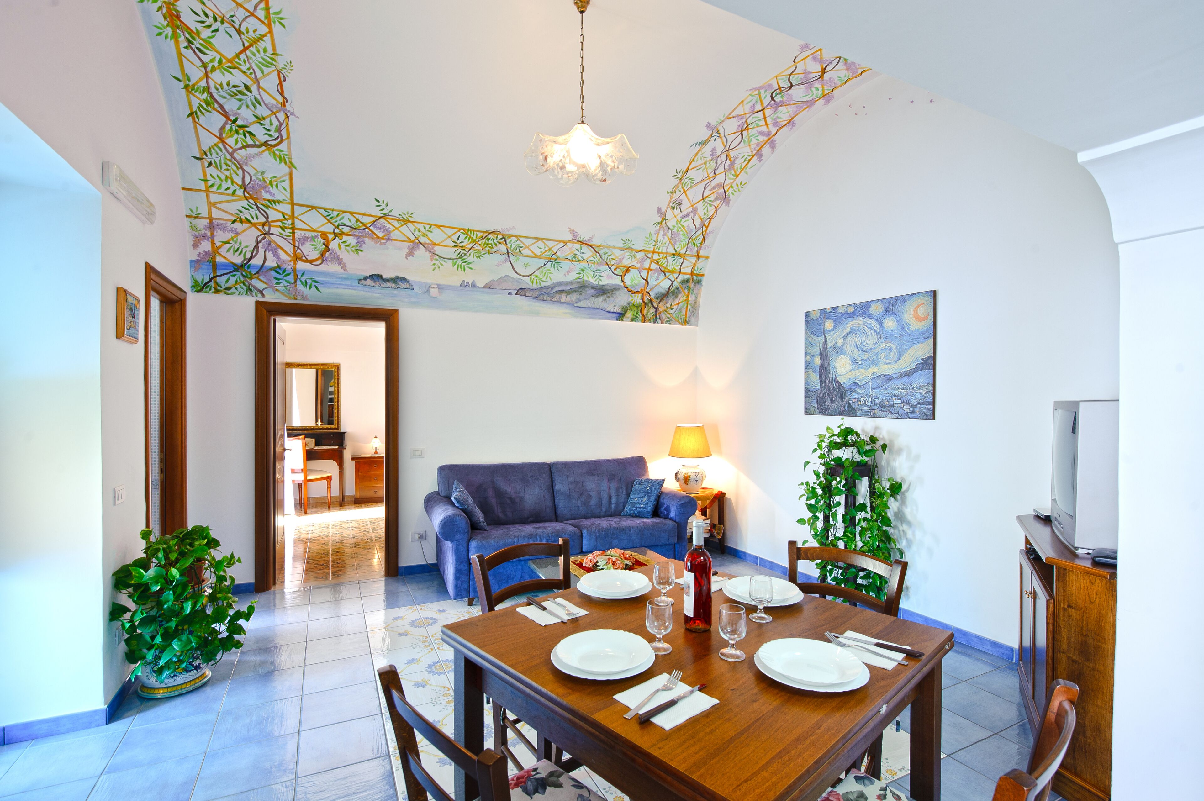 A dining area with a wooden table set for four, a blue sofa, a TV, and a lamp.