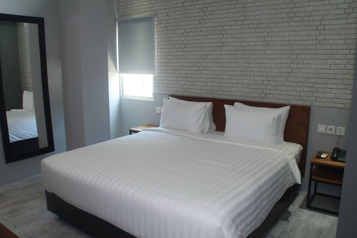 Una cama bien tendida con sábanas blancas en una habitación de hotel, con una pared de ladrillo visto y un espejo grande en un lateral.