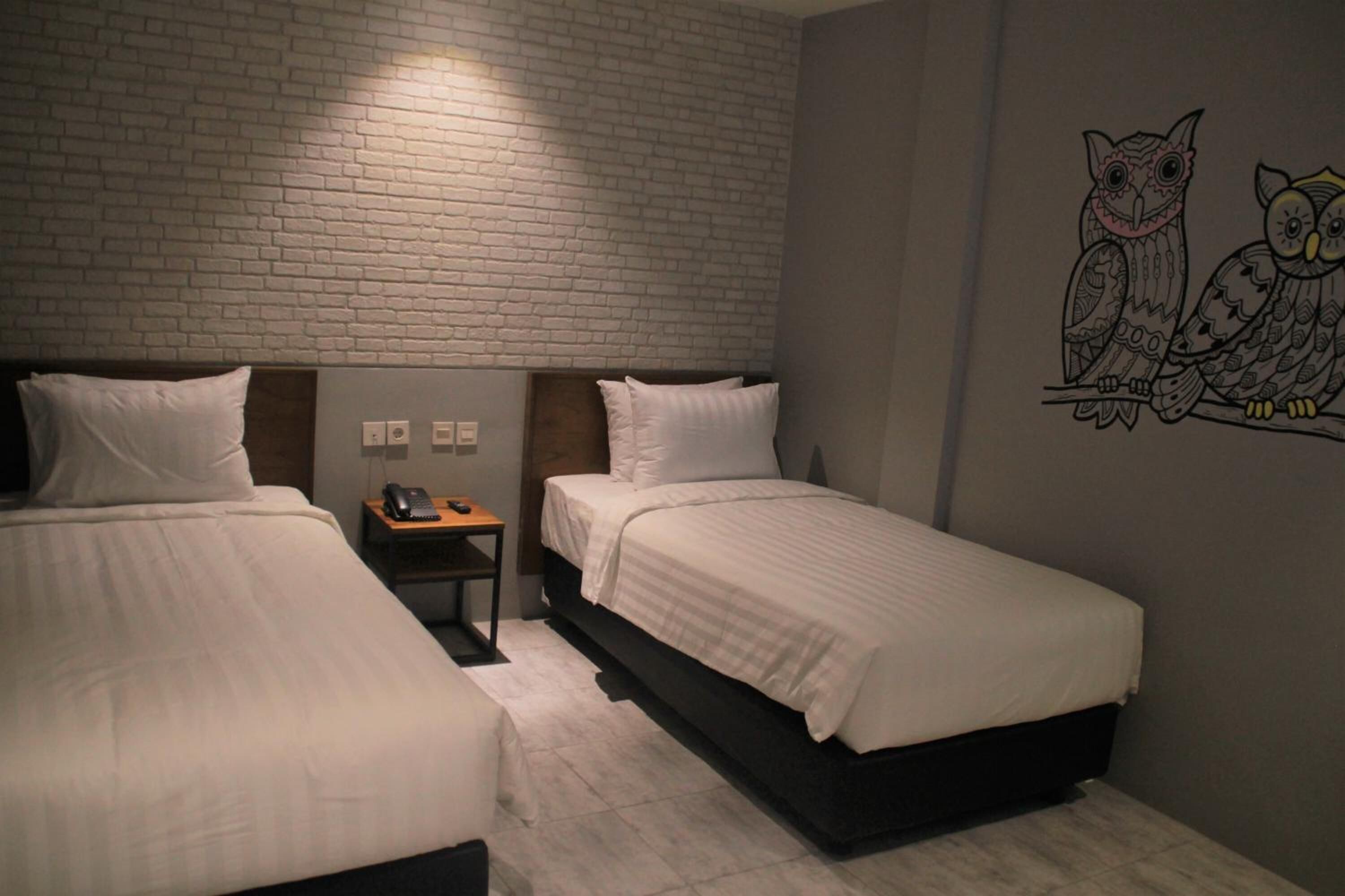 Une chambre d’hôtel avec deux lits simples, une table de chevet avec une lampe et une fresque murale représentant des hiboux.