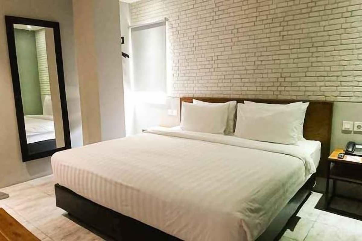 A modern bedroom with a brick wall, a large bed with white bedding, a wooden headboard, and a nightstand with a lamp.