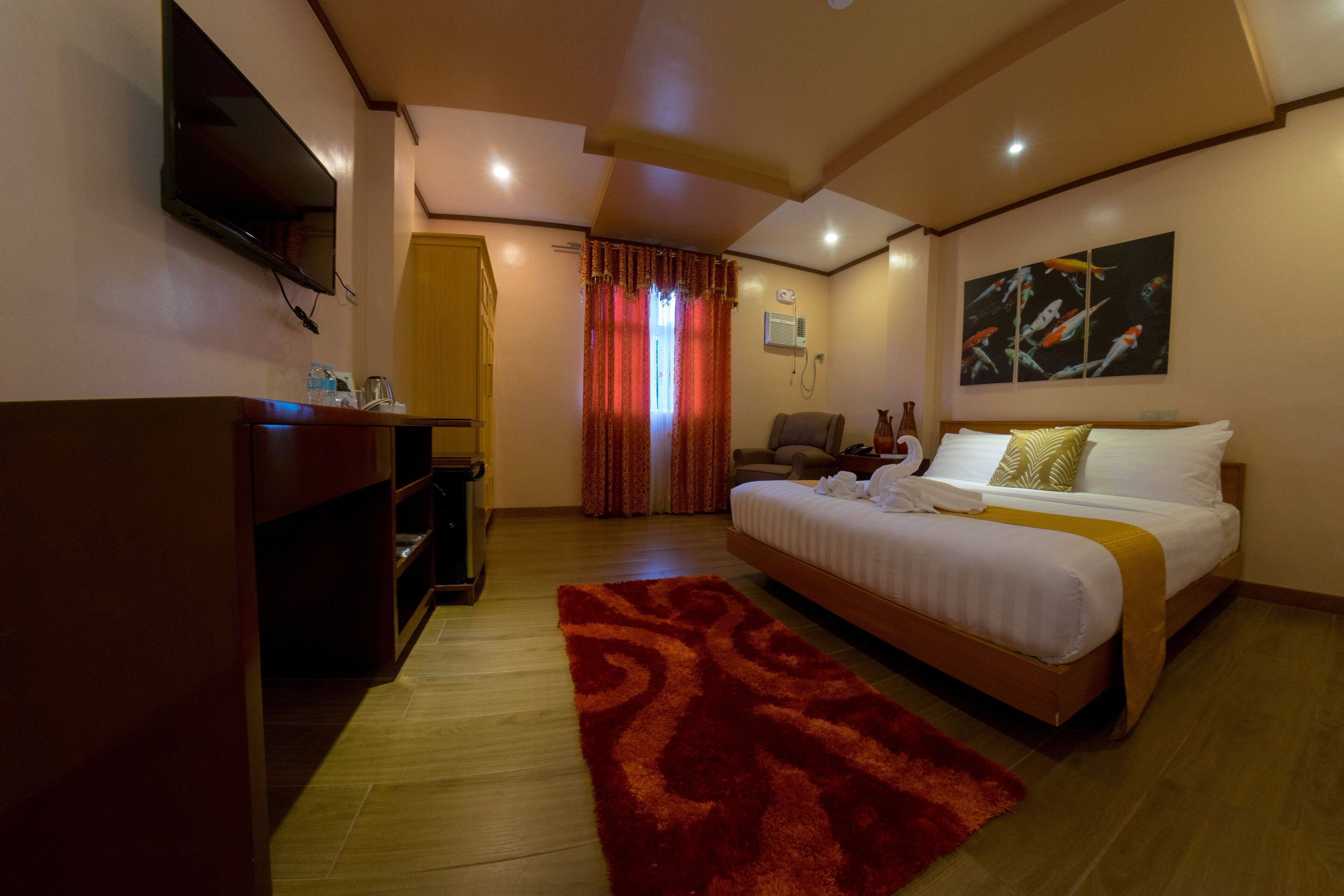 A hotel room with a bed, a TV, a desk, a chair, a red rug, and a painting on the wall.
