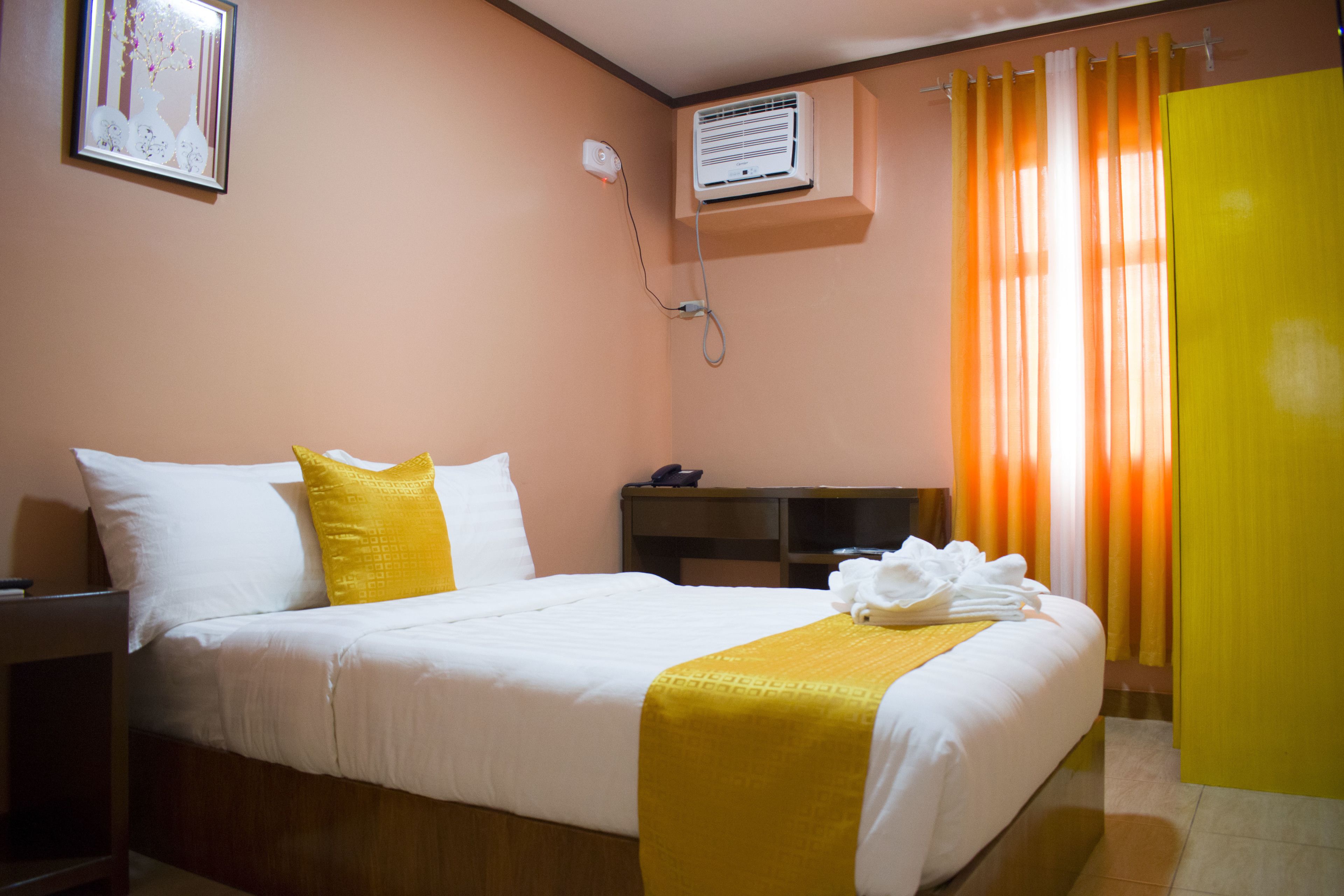 A hotel room with a bed, a bedside table, a telephone, and an air conditioning unit.