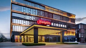 Un hotel moderno con un llamativo letrero de "Hampton", varios pisos y una entrada bien iluminada.