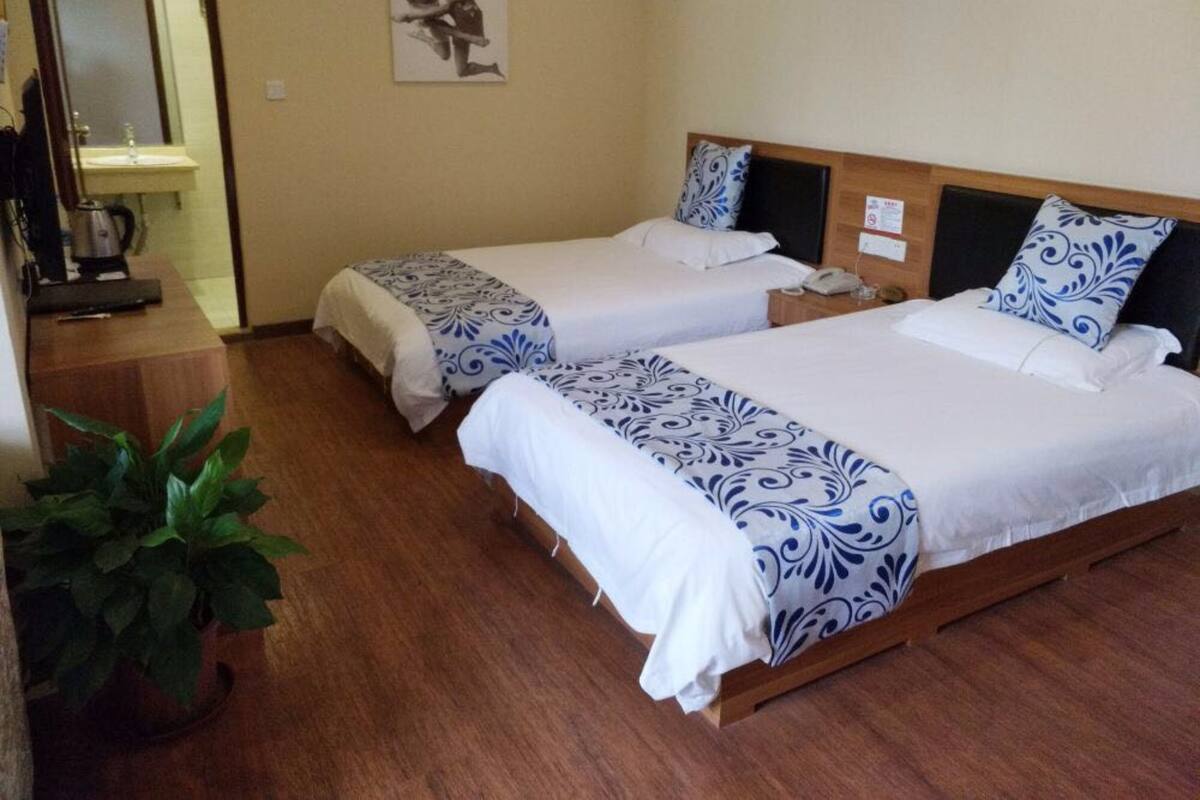A hotel room with two beds, a wooden floor, a potted plant, and a painting on the wall.
