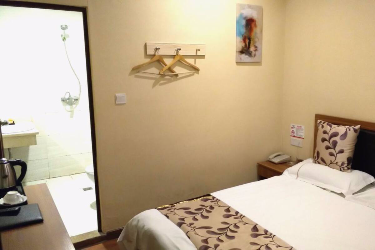 A hotel room with a bed, a bedside table with a phone, a wall-mounted light, and a painting.