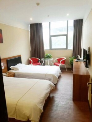 A hotel room with two beds, a desk, a chair, a small table with a plant, and a window with curtains.