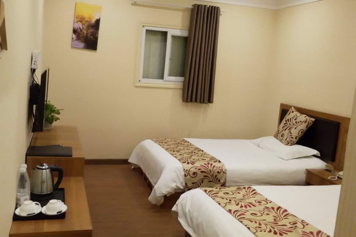 A hotel room with two beds, a small table with a kettle and cups, a window with curtains, and a painting on the wall.
