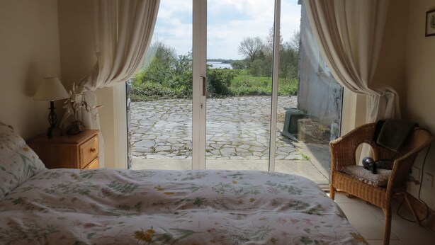 A bedroom with a bed, a nightstand, a wicker chair, and a view of a garden through a glass door. - Lakeside Country Cottage. 
 (Headford)