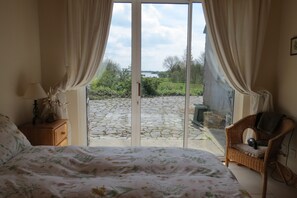 A bedroom with a bed, a nightstand, a wicker chair, and a view of a garden through a glass door. - Lakeside Country Cottage. 
 (Headford)