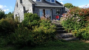 A stone house with a sloped roof, a front porch, and a garden with various plants and flowers. - Lakeside Country Cottage. 
 (Headford)